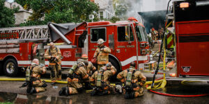A group of firefighters behind a firetruck drinking water and cooling off, as other firefighters enter a building with smoke