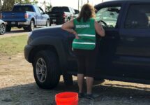 Woman wearing a Green Cross reflective vest stands next to a vehicle and talks to the driver
